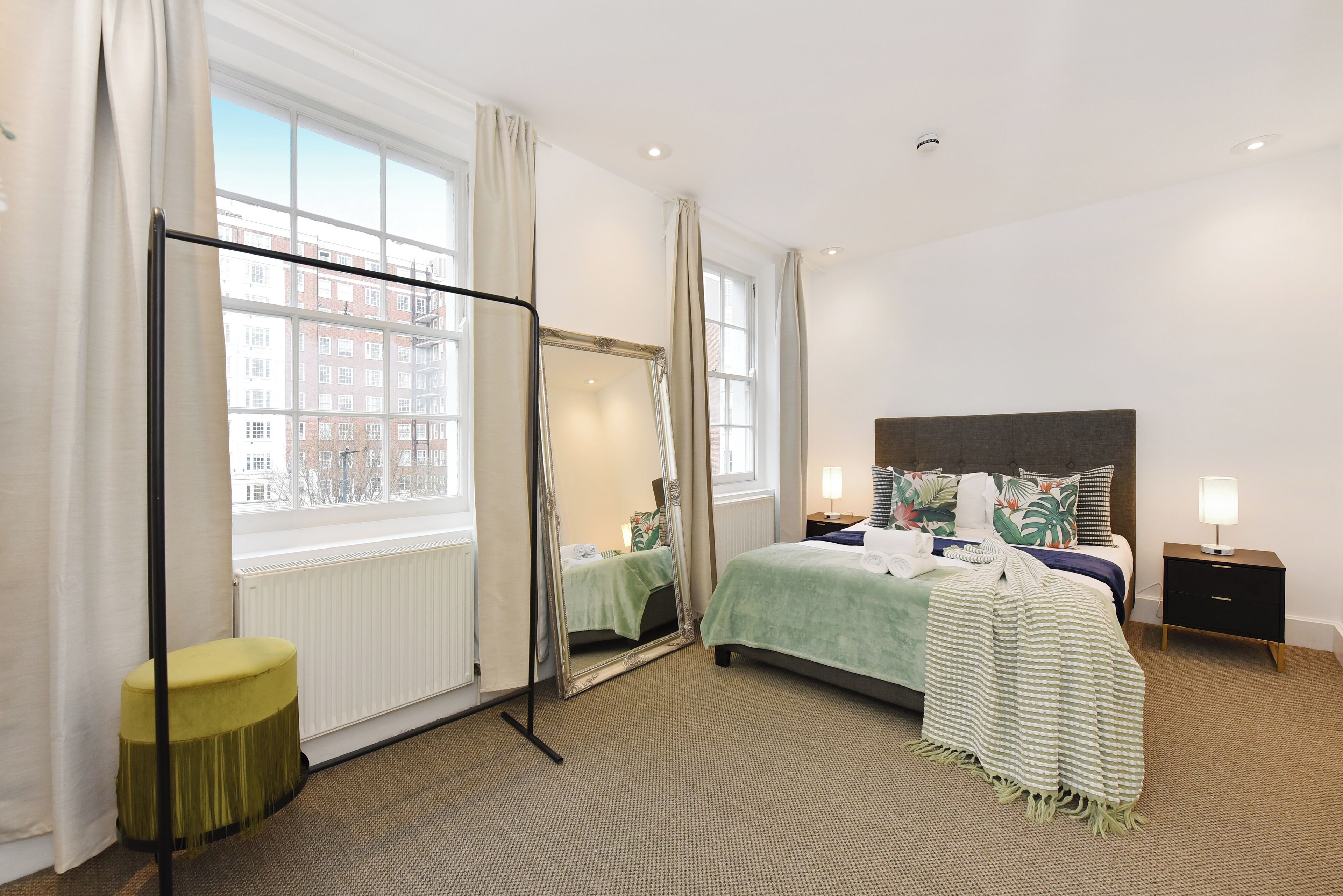 A hotel room with a large window, a bed with a green bedspread, a nightstand with a lamp, and a mirror.