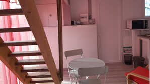 A room with a wooden staircase, a white table with chairs, a white door, a microwave on a shelf, and a trash bin.
