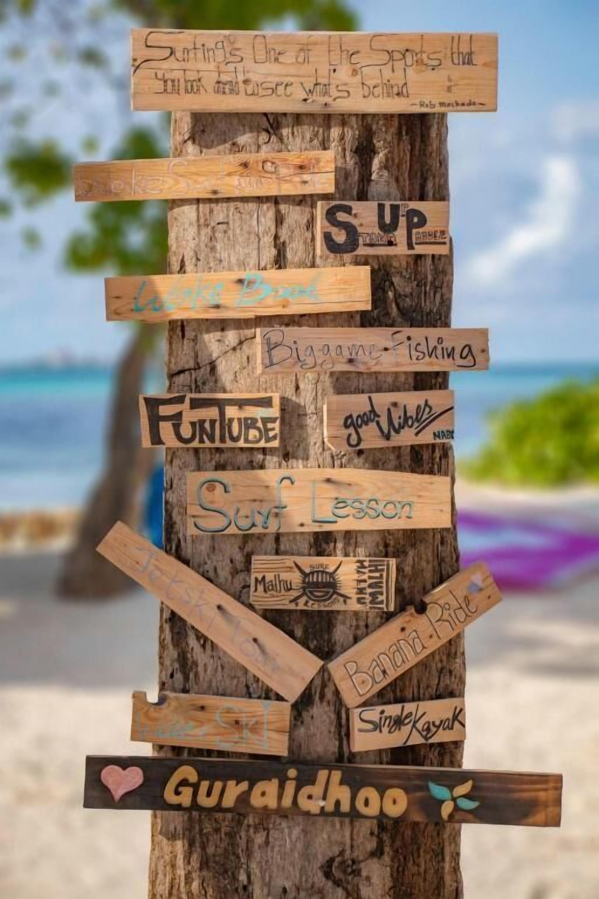 A wooden signpost with various phrases and logos, such as "Surf Lesson" and "Gurai dhoo," against a beach backdrop.