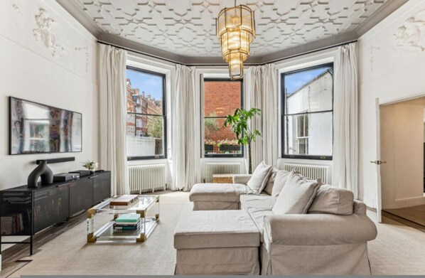 A modern living room with a sectional sofa, a coffee table, and a TV on a black stand. There are large windows with curtains, a radiator, and a plant. - Harley Street Elegance With Private Garden (London)
