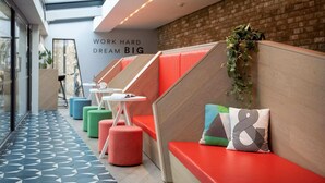 A modern interior with a row of red cushioned seating areas, a brick wall, and a geometric patterned floor. - For Students Only - Studios in Cambridge (Cambridge)