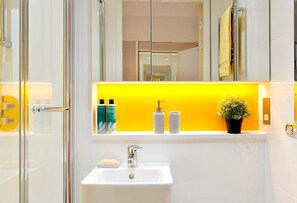 Ein modernes Badezimmer mit einem weißen Waschbecken, einer Duschkabine aus Glas und einem gelblich beleuchteten Bereich mit Pflegeprodukten.