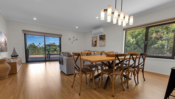 A spacious dining area with a wooden table and chairs, a sofa, and a large window offering a view of the outdoors. - Anira: A Stylish New Escape in Lakes Entrance (Lakes Entrance)