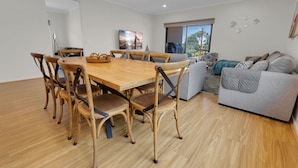 A modern living space with a wooden dining table, wicker chairs, a grey sofa, and a large window with a view. - Anira: A Stylish New Escape in Lakes Entrance (Lakes Entrance)