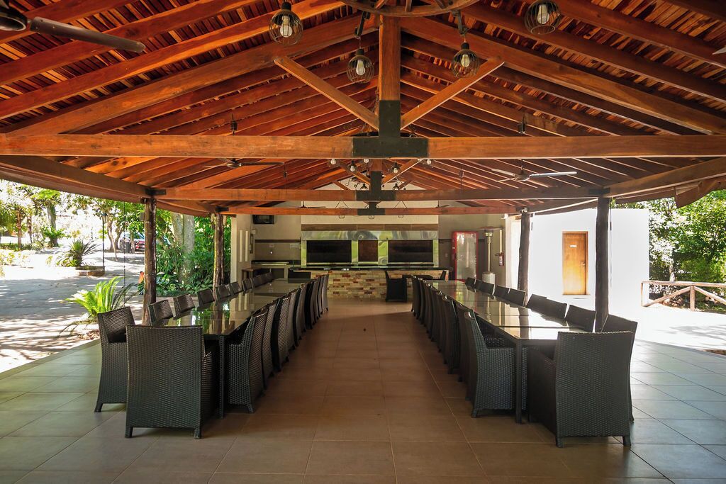 A covered outdoor dining area with a long table and chairs, a stone wall, and a view of a building in the background.