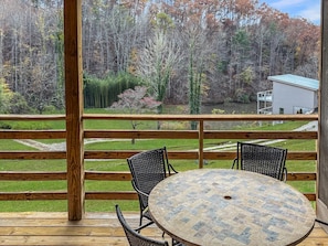 A wooden deck with a round table and two chairs, overlooking a forested area with a house in the background. - King suite in Nantahala Village w/ fireplace, WiFi, and mtn views. Free train, rafting, and zip tix (Bryson City)