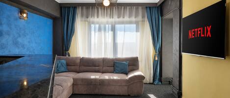 Een moderne woonkamer met een hoekbank, een flatscreen televisie en een groot raam met dunne gordijnen.