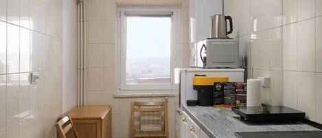 A compact kitchen with white cabinets, a wooden chair, a window, and a countertop with a kettle and a coffee maker.
