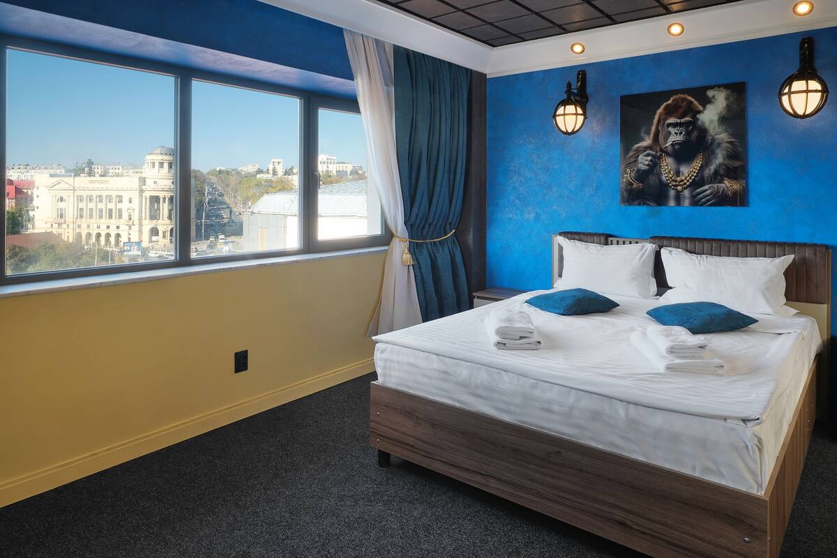 A hotel room with a large bed, a view of a cityscape, and two wall-mounted lights.