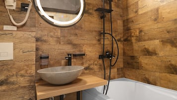 A modern bathroom with a wooden wall, a round mirror, a vessel sink, a handheld showerhead, and a white bathtub.