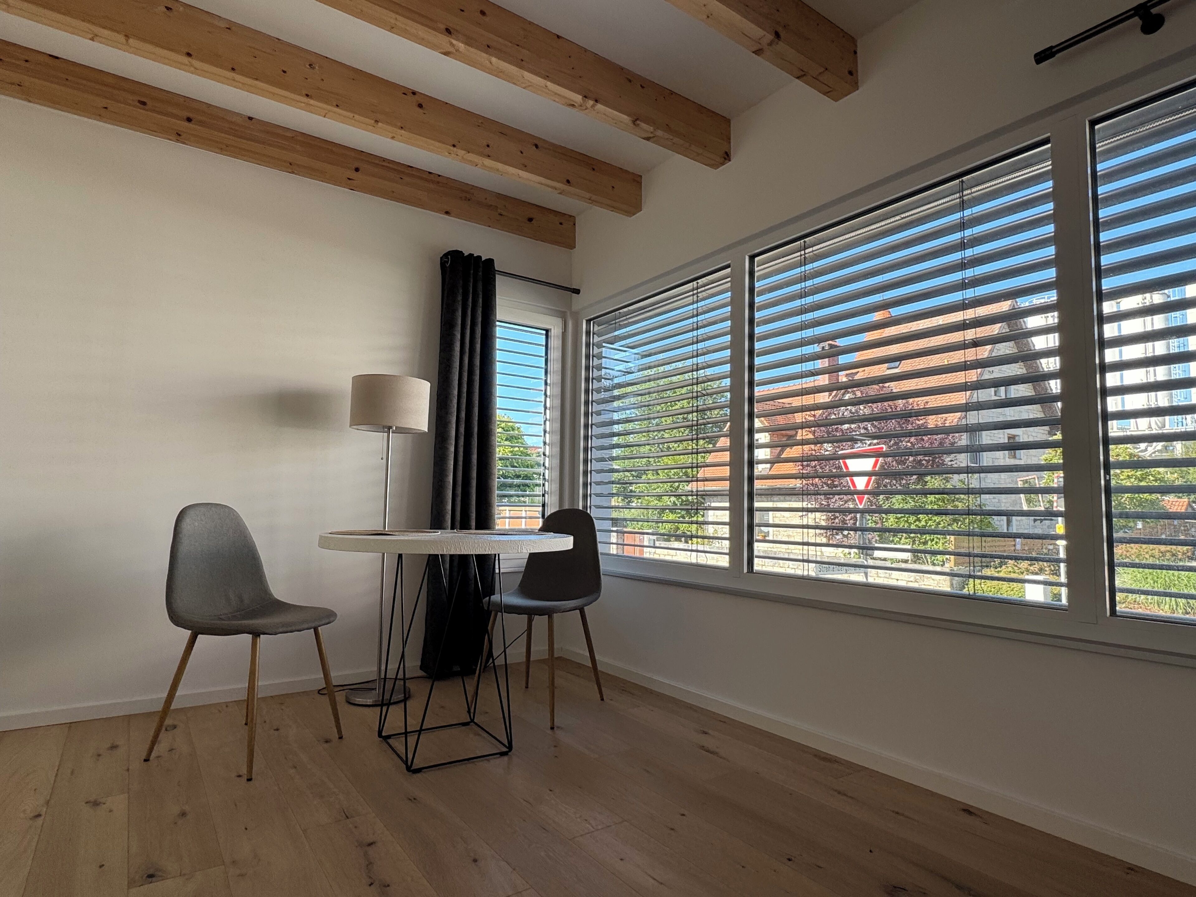 Ein Zimmer mit Tisch, zwei Stühlen, einer Lampe und großen Fenstern mit Jalousien.
