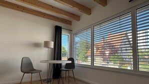 Una habitación con una mesa, dos sillas, una lámpara y ventanas grandes con persianas.