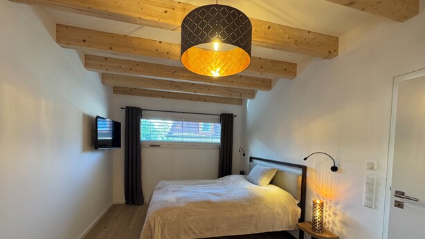 Un dormitorio con una cama, una lámpara colgante, una ventana con cortinas y un televisor en la pared.