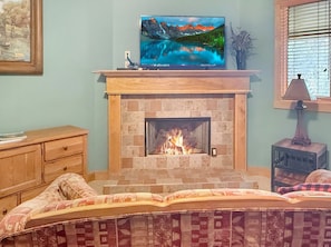 A room with a fireplace, a television mounted above it, a bed with patterned bedding, and a window with blinds. - Nantahala Village king suite w/ fireplace, WiFi, and mtn views. Free train, rafting, and zip tix (Bryson City)