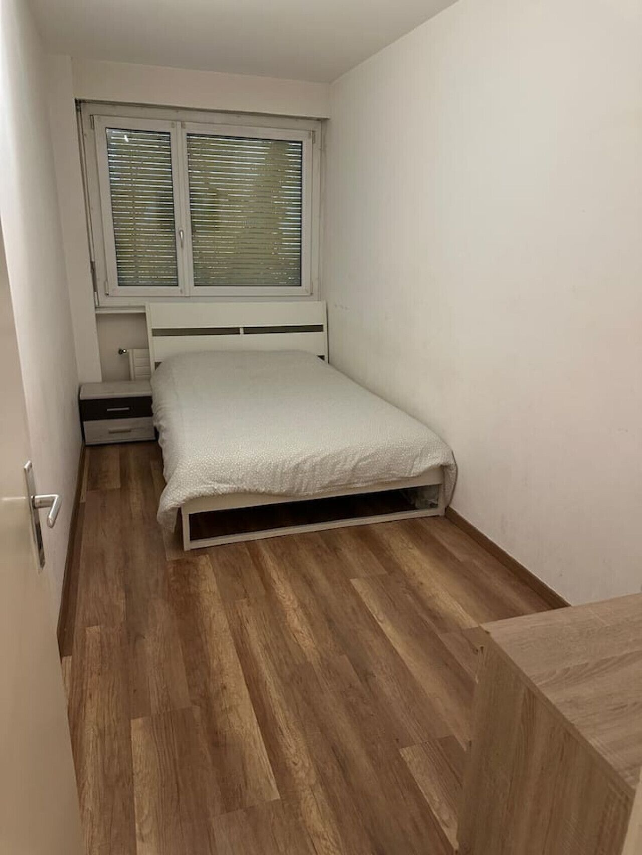 Ein kleines Zimmer mit einem Einzelbett, Holzboden, einem Fenster mit Jalousien und einem Nachttisch.