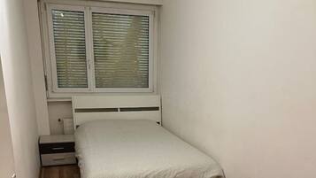 Ein kleines Zimmer mit einem Einzelbett, Holzboden, einem Fenster mit Jalousien und einem Nachttisch.