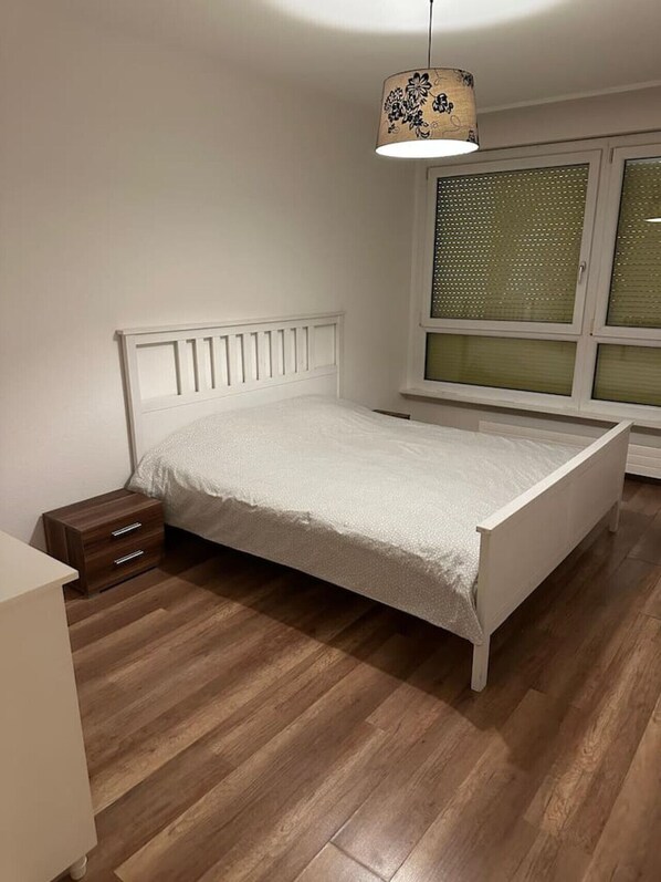 Une chambre avec un plancher en bois, un lit blanc avec une tête de lit, une table de chevet, une fenêtre avec des stores et une lampe suspendue.