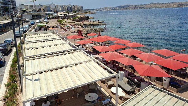 A seaside resort with outdoor seating areas under red umbrellas, overlooking the sea. - Steps Away from the Sea- Beach Front Free Parking! (San Pawl il-Baħar)