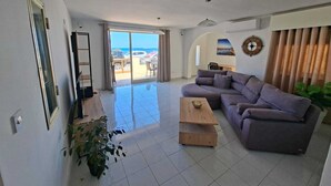 A spacious living room with a large sliding glass door leading to a balcony, a grey sectional sofa, a wooden coffee table, and a potted plant. - Steps Away from the Sea- Beach Front Free Parking! (San Pawl il-Baħar)