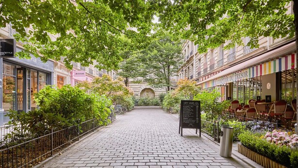 En brostensbelagt stræde med butikker på hver side, en midterste arkade og udendørs siddepladser med et menuskilt.