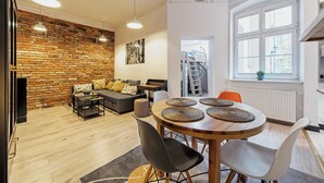 A modern living room with a brick wall, a sofa, a dining table with woven placemats, and a ladder leading to a loft. - RentPlanet - Wyspiańskiego (Poznań)