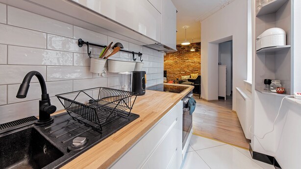 A modern kitchen with white cabinetry, a black sink, and a wooden countertop. There is a brick wall in the background and a hallway leading to another room. - RentPlanet - Wyspiańskiego (Poznań)