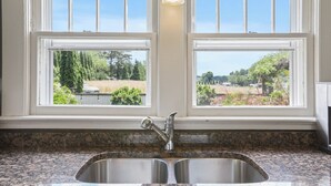Cozinha com bancada de granito, pia de aço inoxidável e duas janelas com vista para uma paisagem rural.
