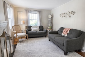 A cozy living room with a grey sofa, a wooden chair, a small table with a lamp, and a clock on the wall. - Peaceful Blue Ridge Farmhouse- Mountain Views,Hot Tub, Fire Pit. Pet Friendly (Candler)