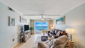 Ett vardagsrum med en soffa, ett soffbord och en TV, en takfläkt och utsikt över havet genom stora fönster.
