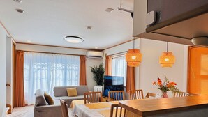 A modern living room with a sofa, dining table, and chairs. There is a TV on a stand, a potted plant, and a window with curtains. - Kamei House (Tokyo)