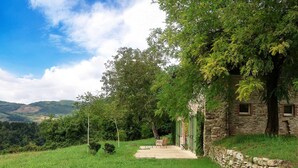 Ein Steinhaus mit überdachter Terrasse, umgeben von üppiger Vegetation und Hügeln im Hintergrund.