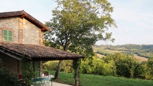 Ein rustikales Steinhaus mit Ziegeldach, einer überdachten Terrasse mit Gartenmöbeln und Blick auf sanfte Hügel im Hintergrund.