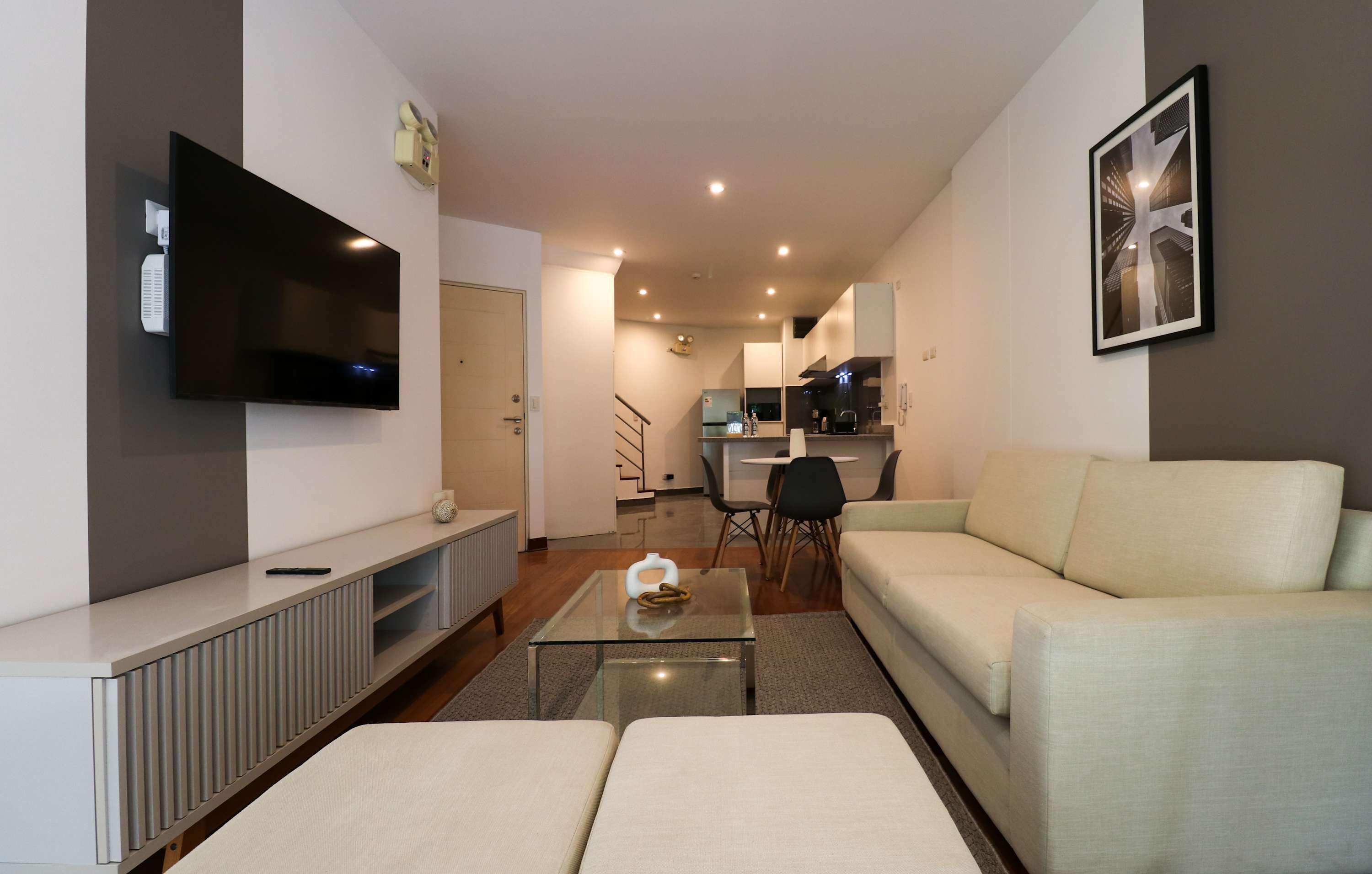 Ein modernes Wohnzimmer mit Flachbildfernseher, Glastisch, Sofa und einem Essbereich im Hintergrund.
