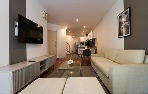 A modern living room with a flat-screen TV, a glass coffee table, a sofa, and a dining area in the background.