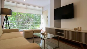 A modern living room with a sofa, a glass coffee table, a TV, and a large window with a view of trees. - w Graceful 1BR With Balcony in Miraflores (Lima)