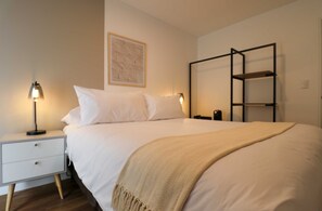Cama bem arrumada com manta bege, luminária de cabeceira, mesinha de cabeceira e prateleira fixada na parede.