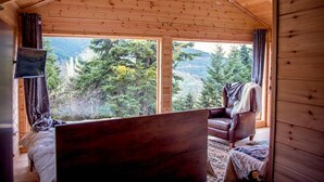 A wooden cabin with a bed, a chair, and a large window offering a view of a forested mountain. - Kedra Village Cozy Chalet - Mountain Getaway (Makrakomi)
