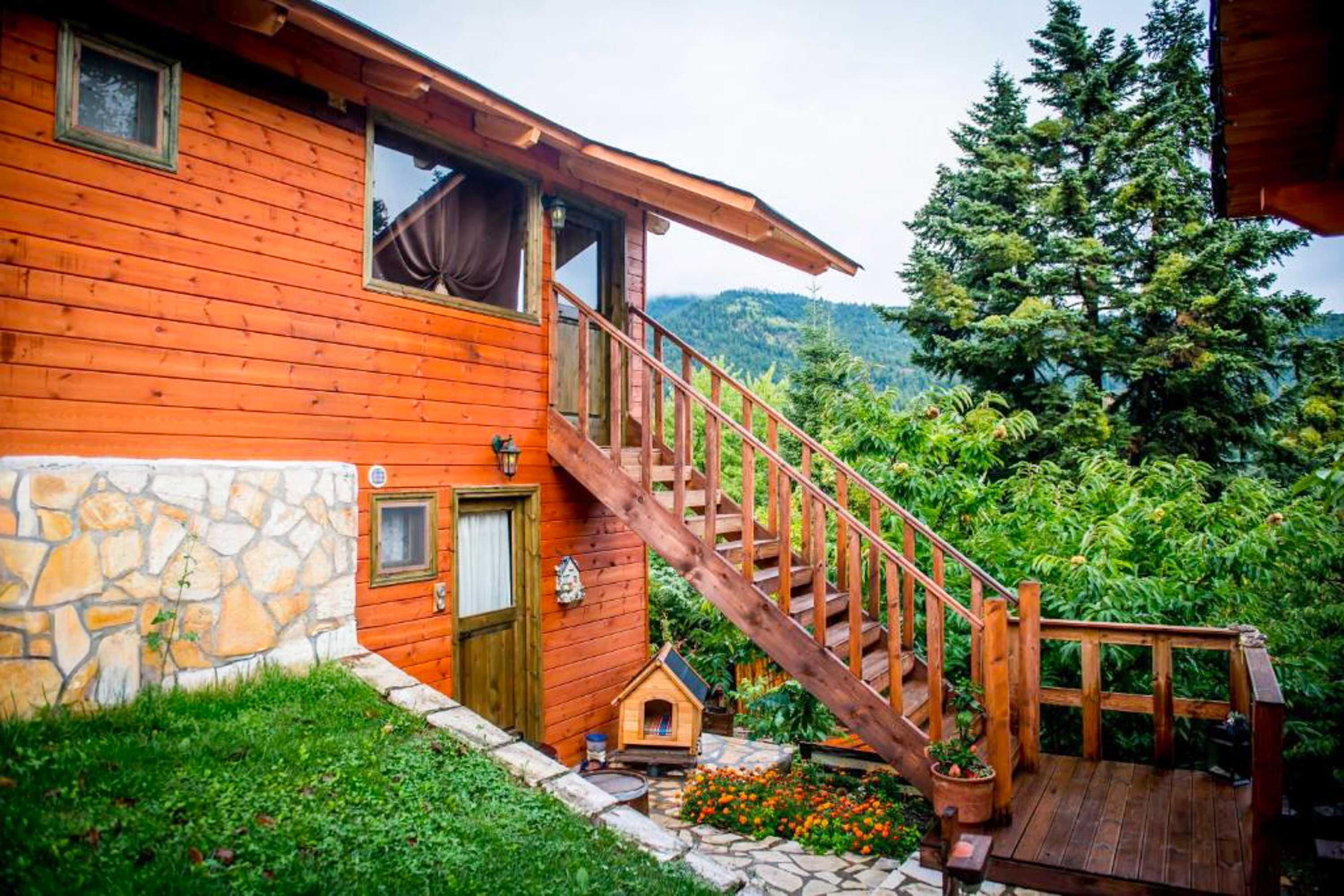 A wooden cabin with a stone wall, a wooden staircase, and a view of a forested mountain.
