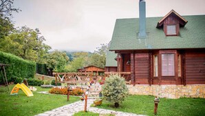A wooden cabin with a stone foundation, a green roof, and a chimney. There is a wooden deck with a swing set and a small garden area with a potted plant and a wheelbarrow. - Kedra Village Superior Chalet - Mountain Escape (Makrakomi)