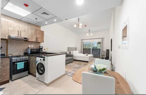 A modern kitchen with a stove, microwave, and washing machine. There is a dining area with a table and chairs, and a sofa in the living room. - Stayshort - Chic Studio Apartment in JVC Sleeps 4 (Dubai)