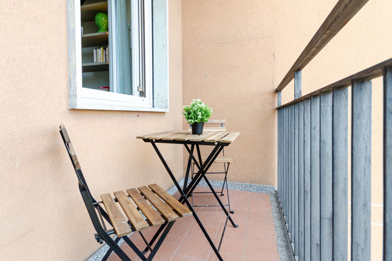 Un pequeño patio al aire libre con mesa y banco de madera, una planta en maceta y una ventana con estanterías.