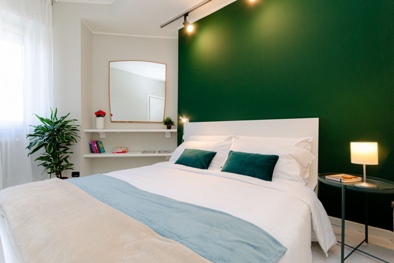 A modern bedroom with a large bed, a green accent wall, a white headboard, and a bedside table with a lamp.