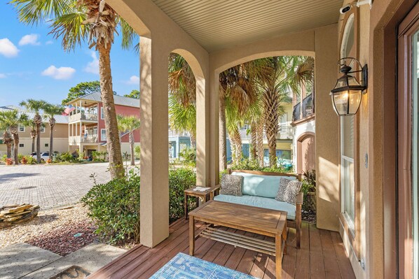 A covered outdoor seating area with a wooden table and sofa, surrounded by palm trees and a view of other buildings. - Sunset Cove: Charming 3BR/3BA Beach House Sleeps 8 with Sunsets & Serenity (Panama City Beach)