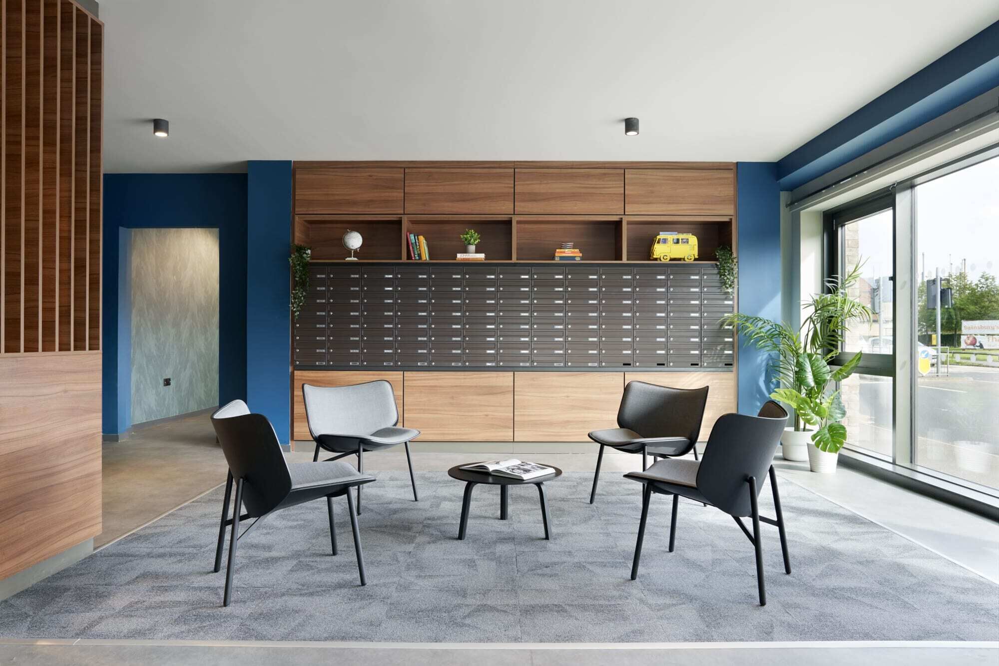 A modern waiting area with a large wall of storage lockers, two black chairs, a small table, and a potted plant.