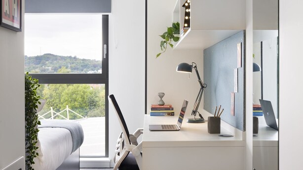 Et soveværelse med en seng, et skrivebord med en bærbar computer, en skrivebordslampe og et spejl. Der er et vindue med udsigt til grønt og et bjerg.