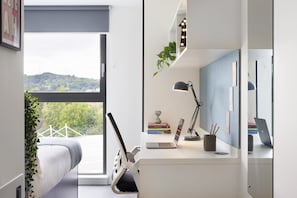 A bedroom with a bed, a desk with a laptop, a desk lamp, and a mirror. There is a window with a view of greenery and a mountain. - For Students Only Ensuites in Edinburgh (Edinburgh)