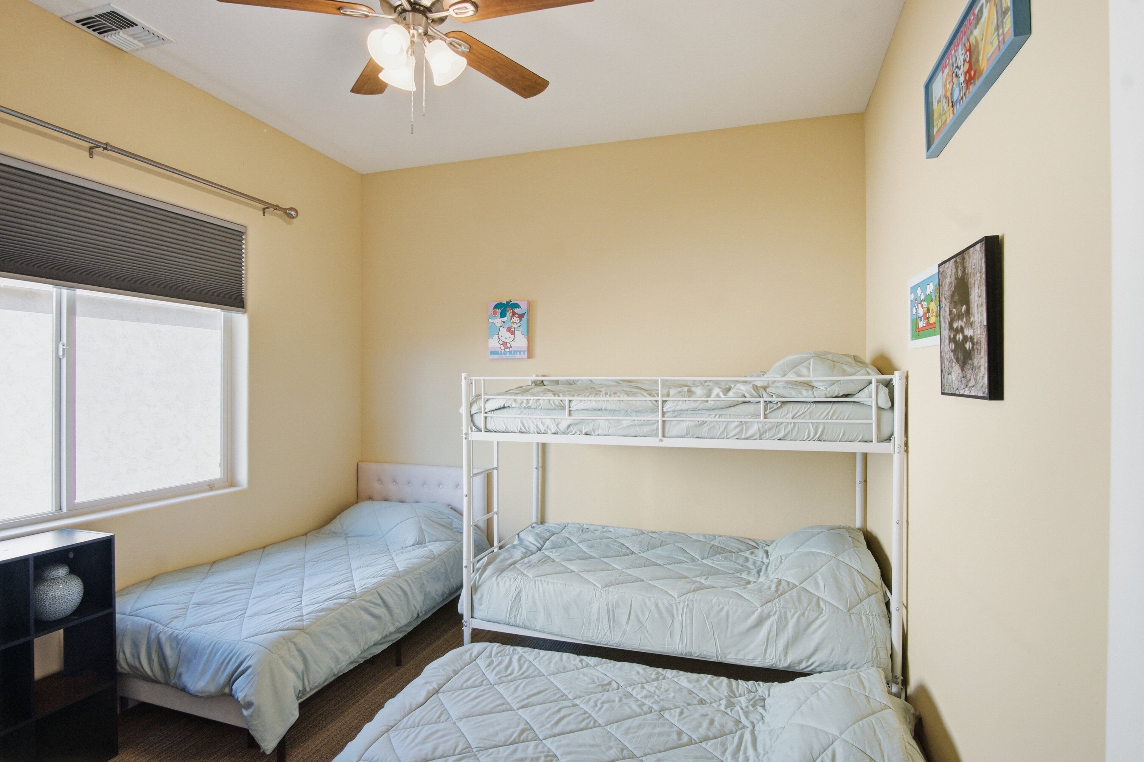 A dormitory room with two bunk beds, a ceiling fan, a window with blinds, and a small desk.