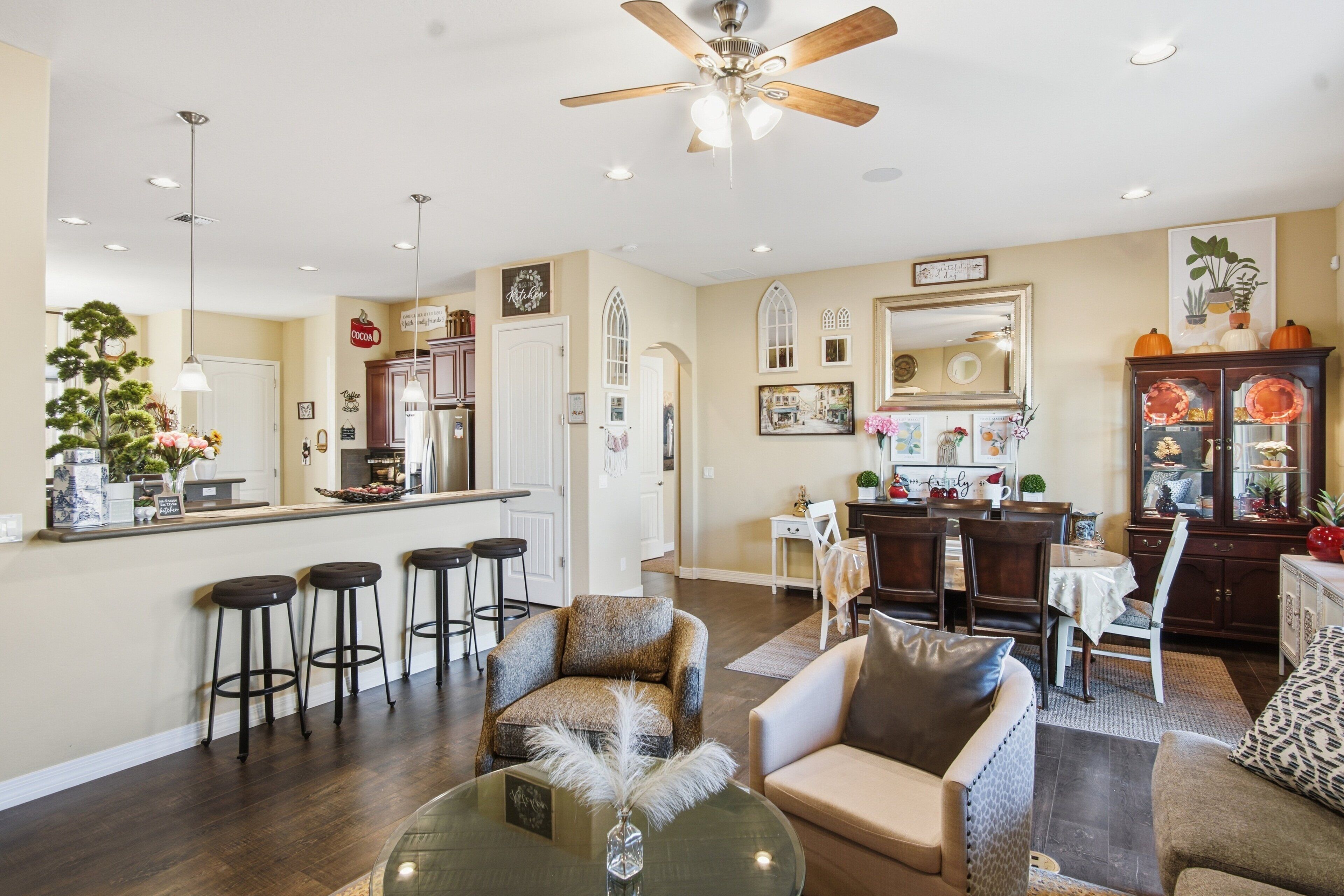 A spacious living room with a high ceiling fan, a dining area with a white table, and a kitchen with a bar counter.
