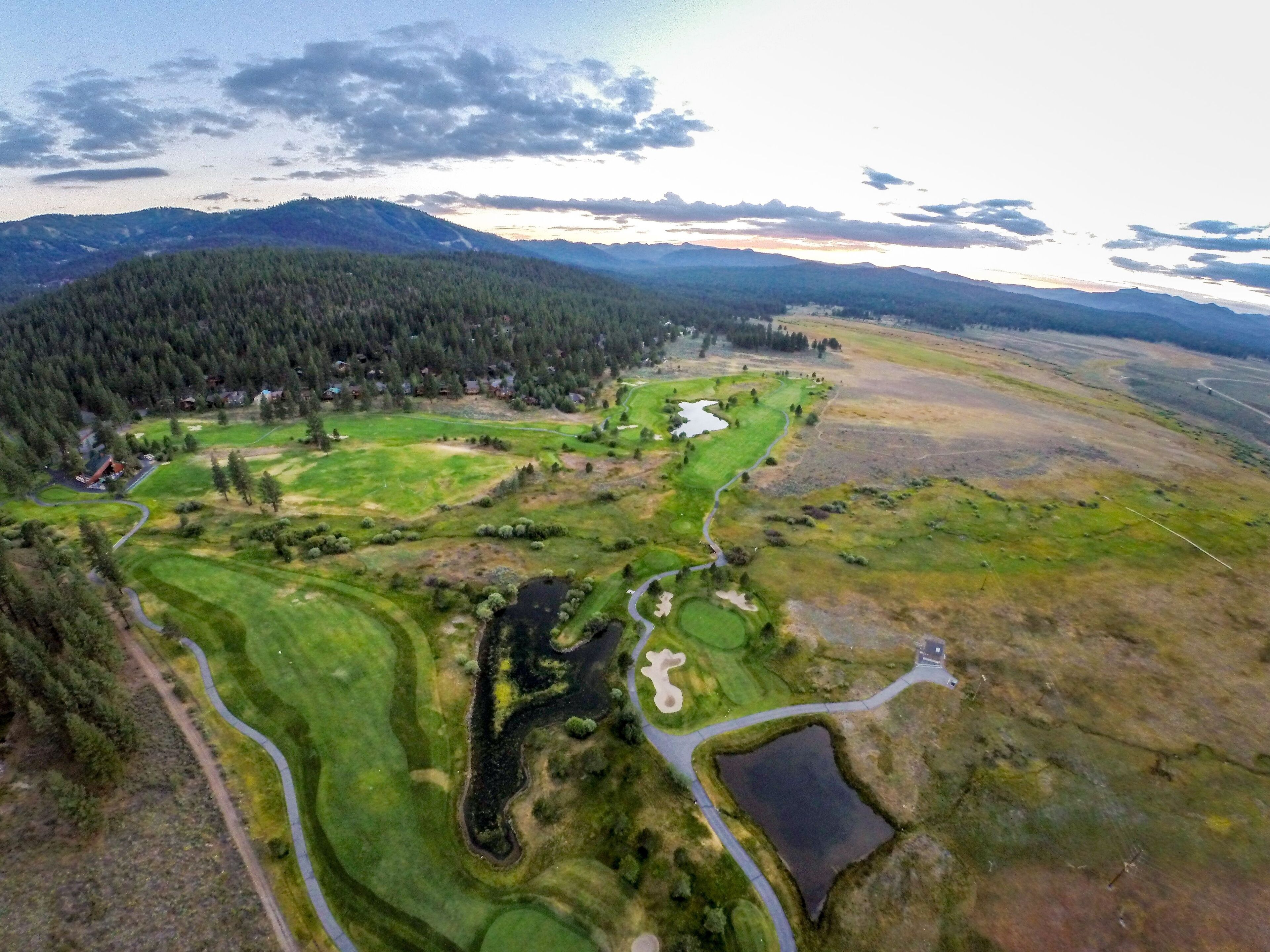 An aerial view of a golf course with multiple greens, water hazards, and a forested area in the background.