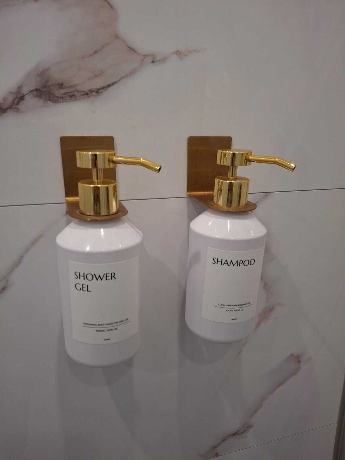 Two white bottles with gold pumps, labeled 'Shower Gel' and 'Shampoo', mounted on a marble wall.